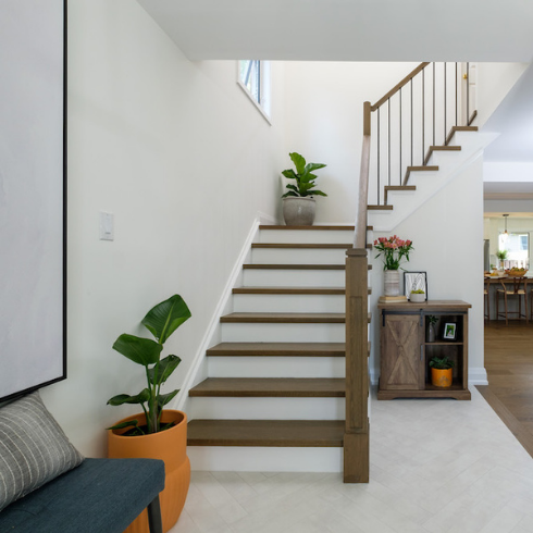 A foyer with a low bench with a large piece of art hung on the wall above it, white herringbone tile floors, wooden steps with white treads, white walls, a rustic wooden farmhouse-style side table and several indoor plants.