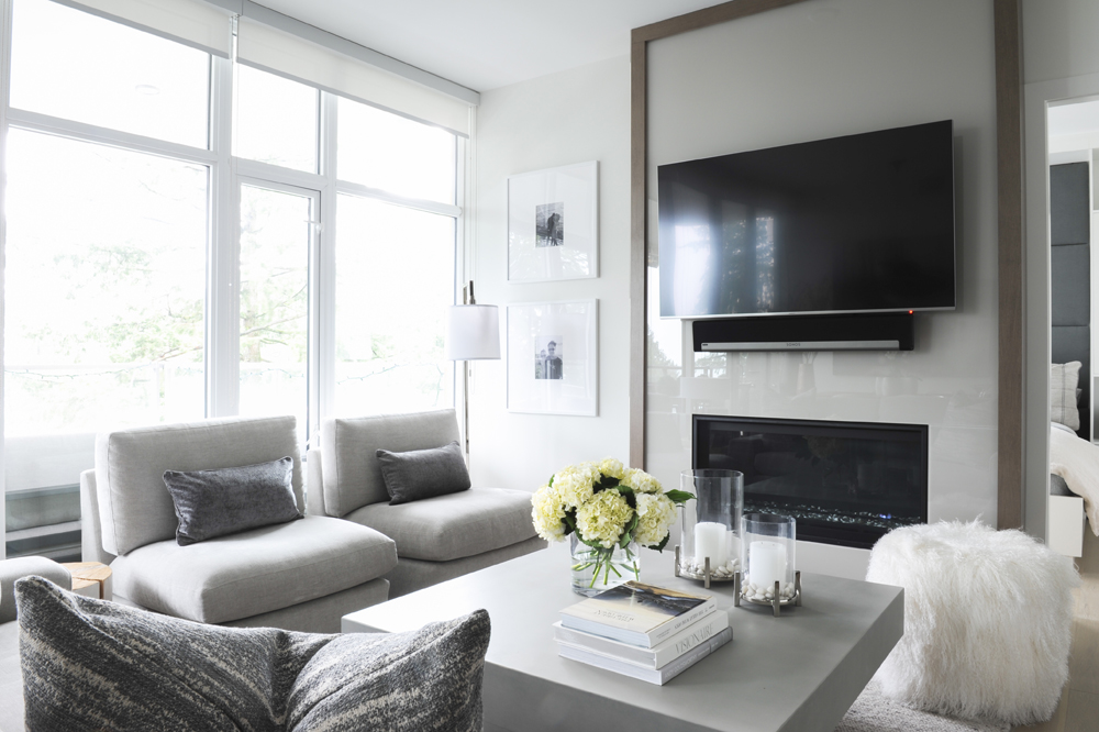 Light filled living room decorated in the transitional style with clean lines and strong furnishings such as two upholstered grey chairs, a square cement coffee table and a large TV positioned over a gas fireplace