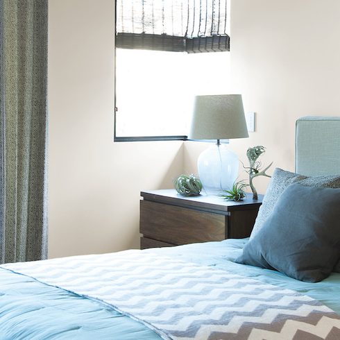 Simple modern bedroom with off white walls painted in BEHR Off White 73, a grey upholstered bed with a blue bedspread and a pile of grey pillows, and two dark wood bedside tables with a glass lamp on each.