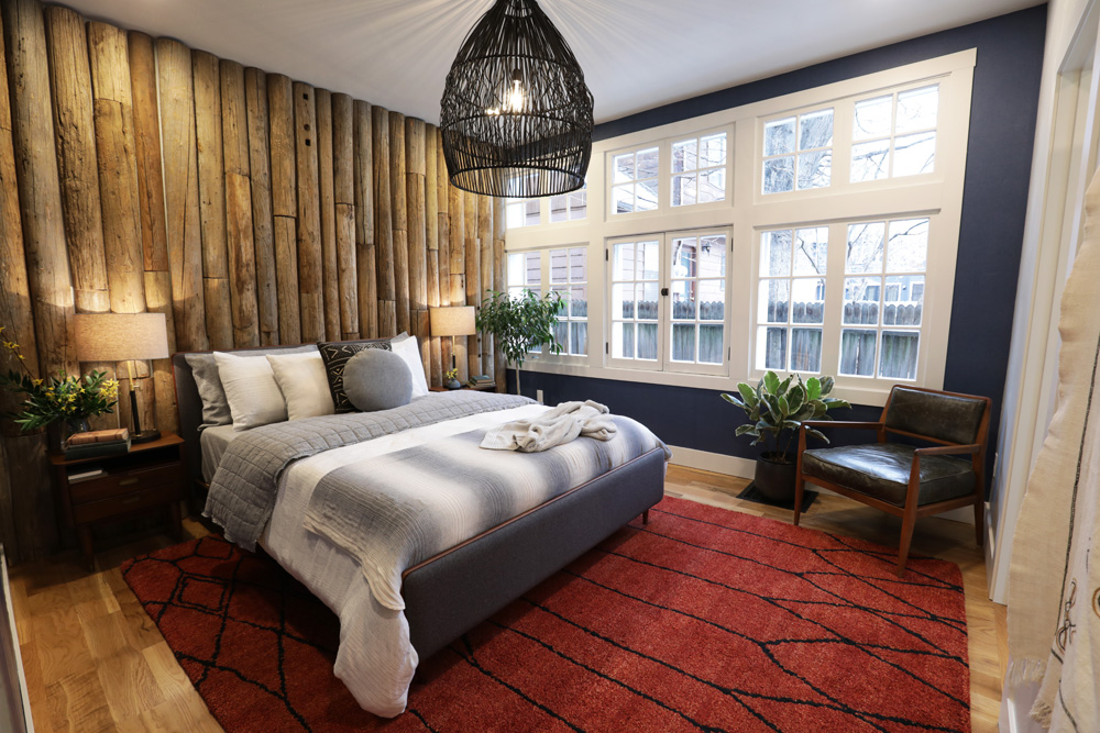 Cozy cabin themed bedroom with a log feature wall behind the grey upholstered bed, large white framed windows, navy walls, a large red run, two bedside tables with laps, a large black wicker pendant lamp, and black leather and wood armchair in the corner designed by Alison Victoria on Home Network’s Rock the Block