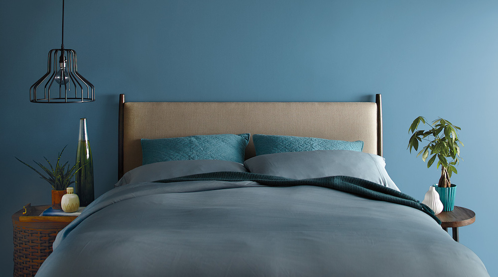 Modern monochromatic bedroom featuring a bed with a tan headboard and blue bedding, a rattan bedside table with a plant and two vases, a wooden bedside table with a plant and vase, an industrial inspired pendant lamp, and walls painted in BEHR Blueprint S470-5