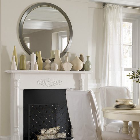 Beautiful fresh dining room with a round white dining table, four white slipcovered dining chairs, a fireplace with a mantle layered with white, yellow and green vases, and walls painted in BERH Yucca White YL-W08 and Ultra Pure White 1850