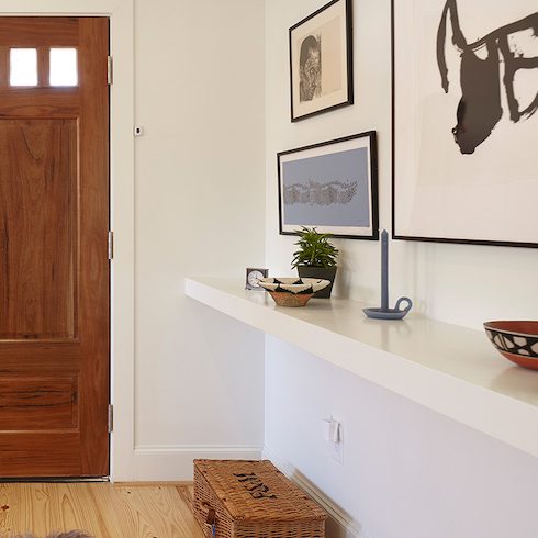 A clean and welcoming entry features a stained wood front door, framed modern art on the walls, a floating shelf with a bowl, plant and candlestick, a potted plant on the floor, and white BEHR Polar Bear 75 paint on the walls.