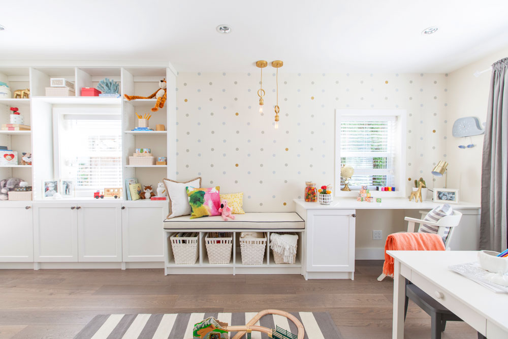 Playroom designed by Jillian Harris on Home Network with built-in wraparound window cubbies with a cushioned seat, quirky gold knotted pendant lamps, white walls, polka dot wallpaper, dark wood floors, and a grey and white striped rug