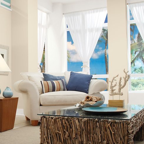 A chic beach-themed living room with a driftwood coffee table, three shell photos on the wall, sheer white drapes, a white couch and blue-striped armchair covered in blue throw pillows, and walls painted in BEHR Roman Plaster PPU7-10, Palais White GR-W15 and Opal Silk PPU12-08