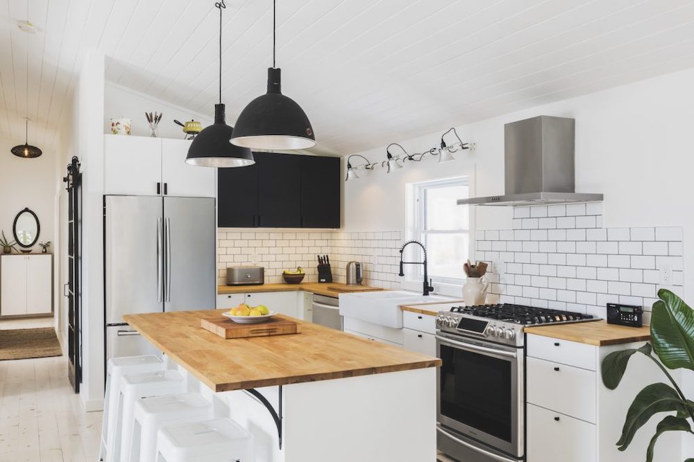 A kitchen with a strong Scandinavian vibe with black grout on the subway tile backsplash, two black island pendants, large wood topped kitchen island, stainless steel appliances and white apron sink