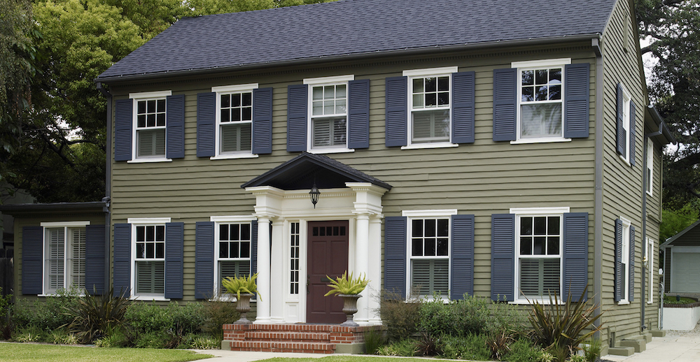 Exterior of a large colonial style house painted in a khaki green BEHR Urban Safari PPU8-04, with white window trim in BEHR White 52, blue shutters in BEHR Poppy Seed PPU15-20 and the burgundy door in BEHR Cherry Cola S130-7