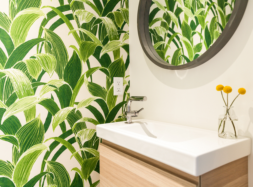 Tropical palm leaf wallpaper pattern in bathroom with a narrow white sink and round black mirror on the wall