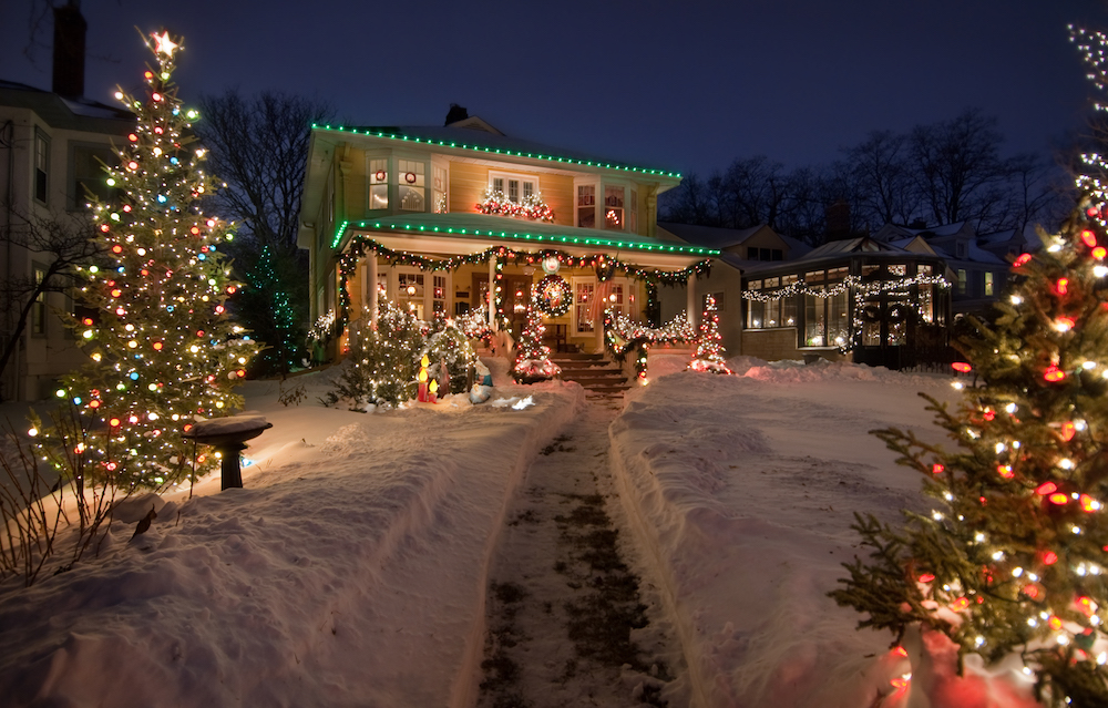 yellow house with green Christmas lights and Christmas trees on snowy front lawn