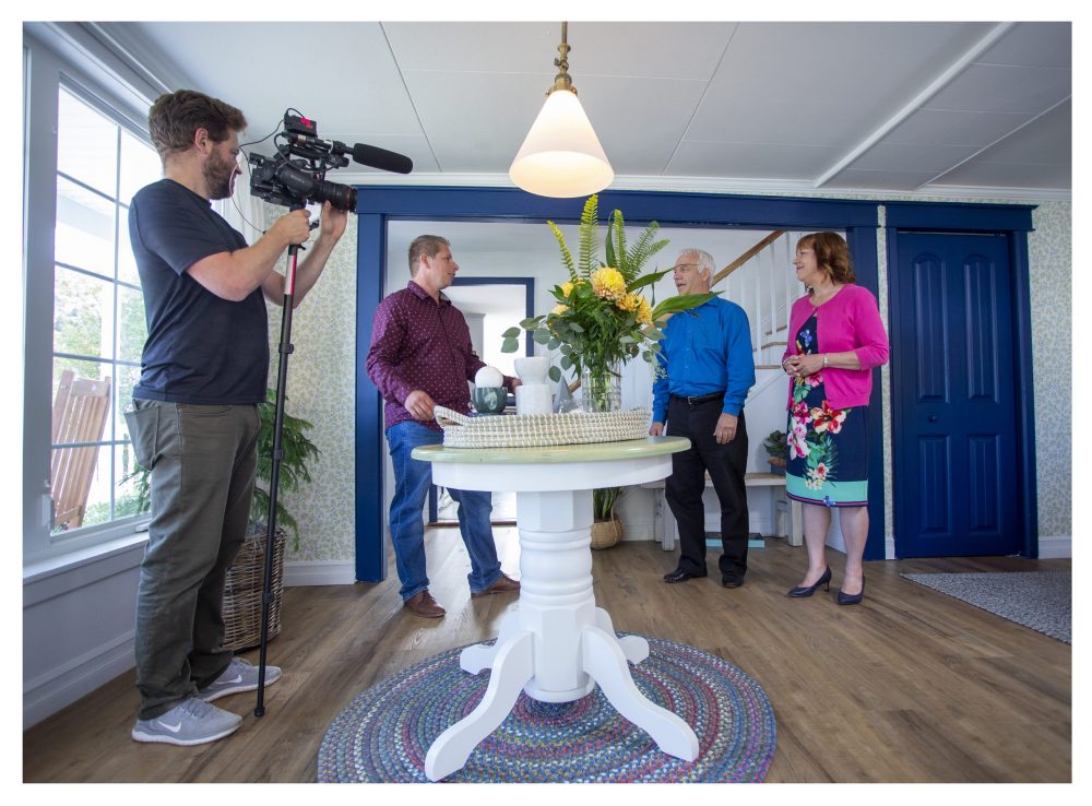 BTS image of a camera man filming Randy and the home owners as they stand in the home’s sunny foyer in front of a small round table