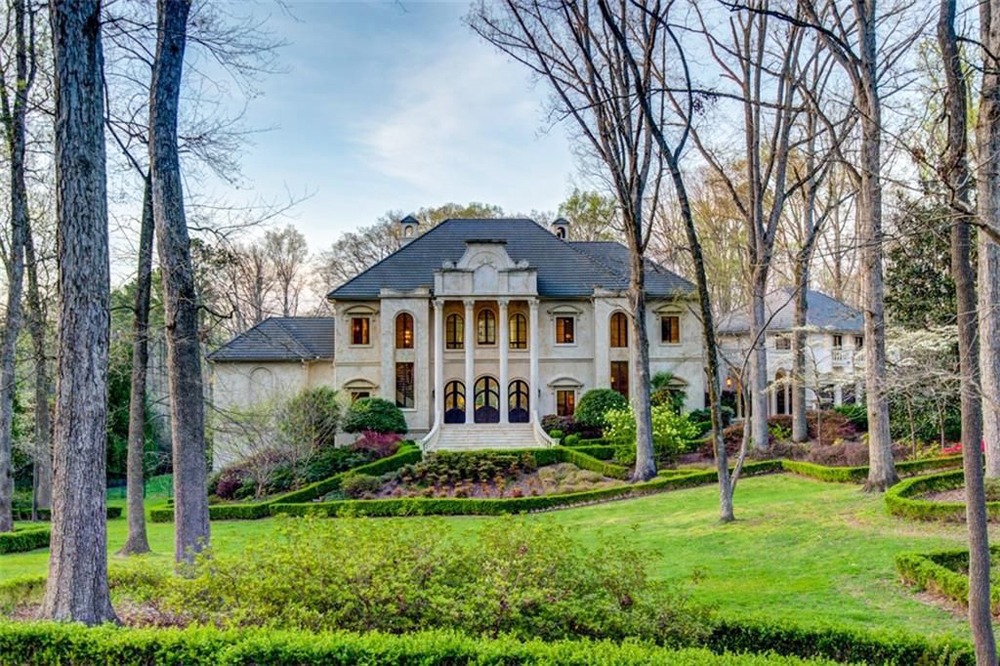 Exterior shot of a mega-mansion on six acres of land, surrounded by a forested area and featuring a sweeping grand staircase leading up to the front door