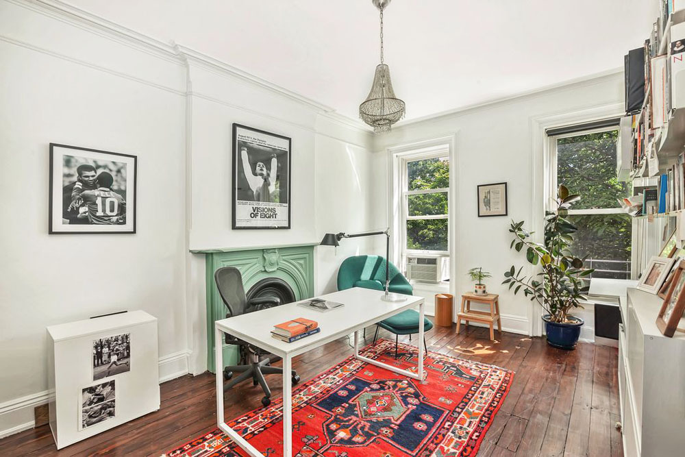 A tiny home office with original wood flooring, area rug, simple white desk and wood-burning fireplace