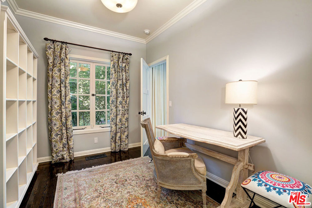 A narrow home office with area rug, patterned curtains and simple wood desk and plenty of bookshelves