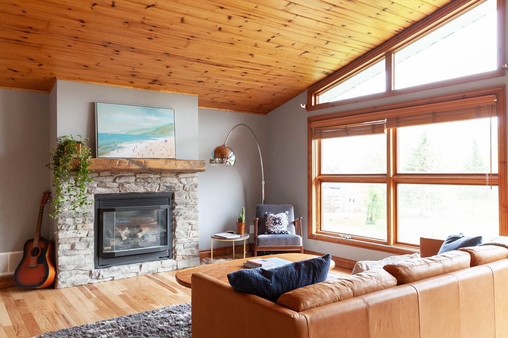 Bright living room with wood ceiling and stone fireplace. A plant with trailing vines sits on the fireplace mantle.