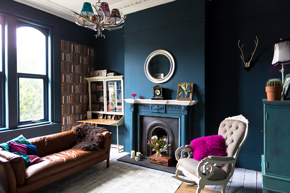 Living room with deep blue walls and fireplace. In front of the fireplace is a bouquet of flowers in a vase on top of a vintage suitcase.