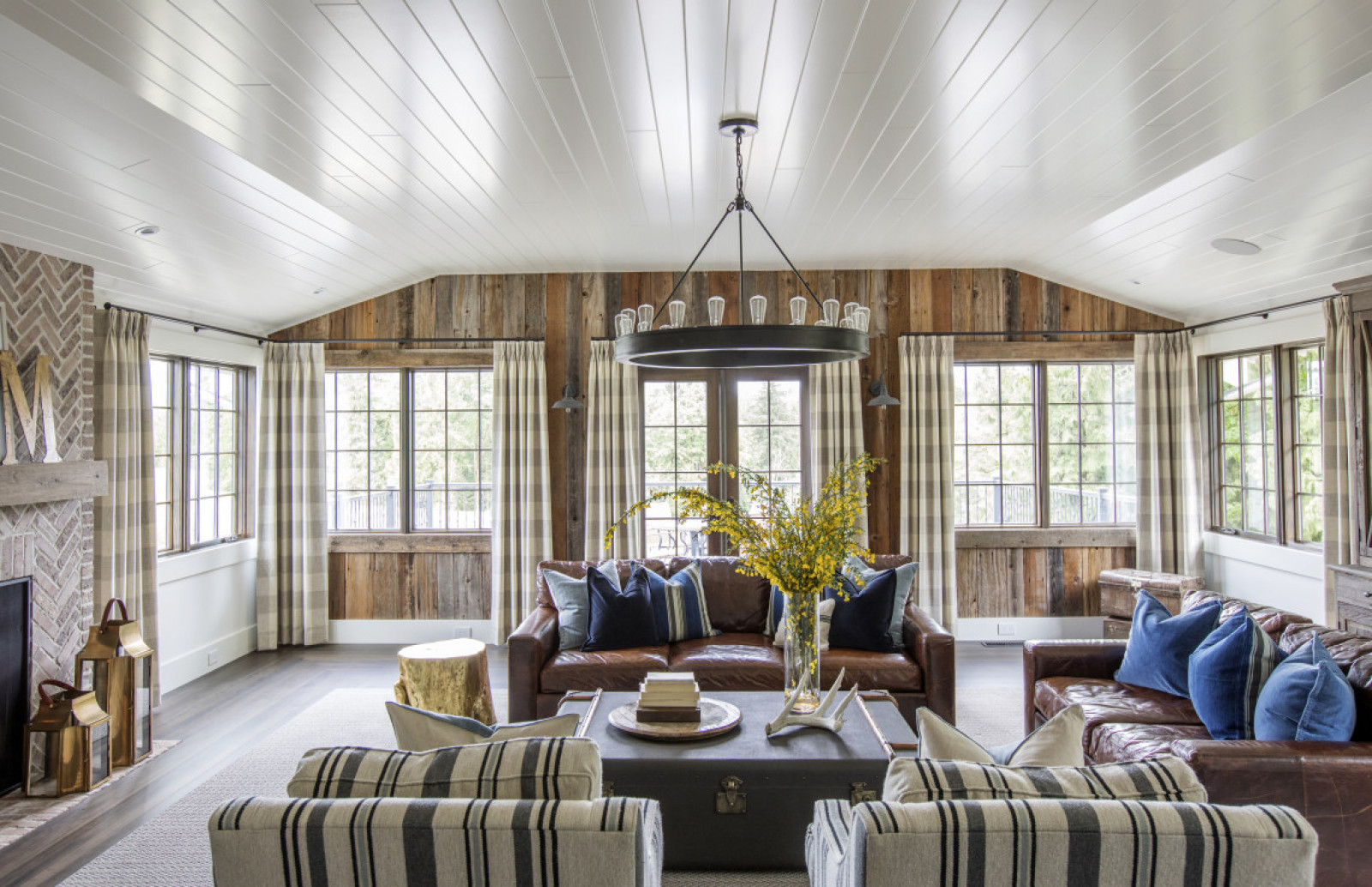 living room with white wood ceiling, wood plank walls, windows, two brown leather sofas, two striped arm chairs