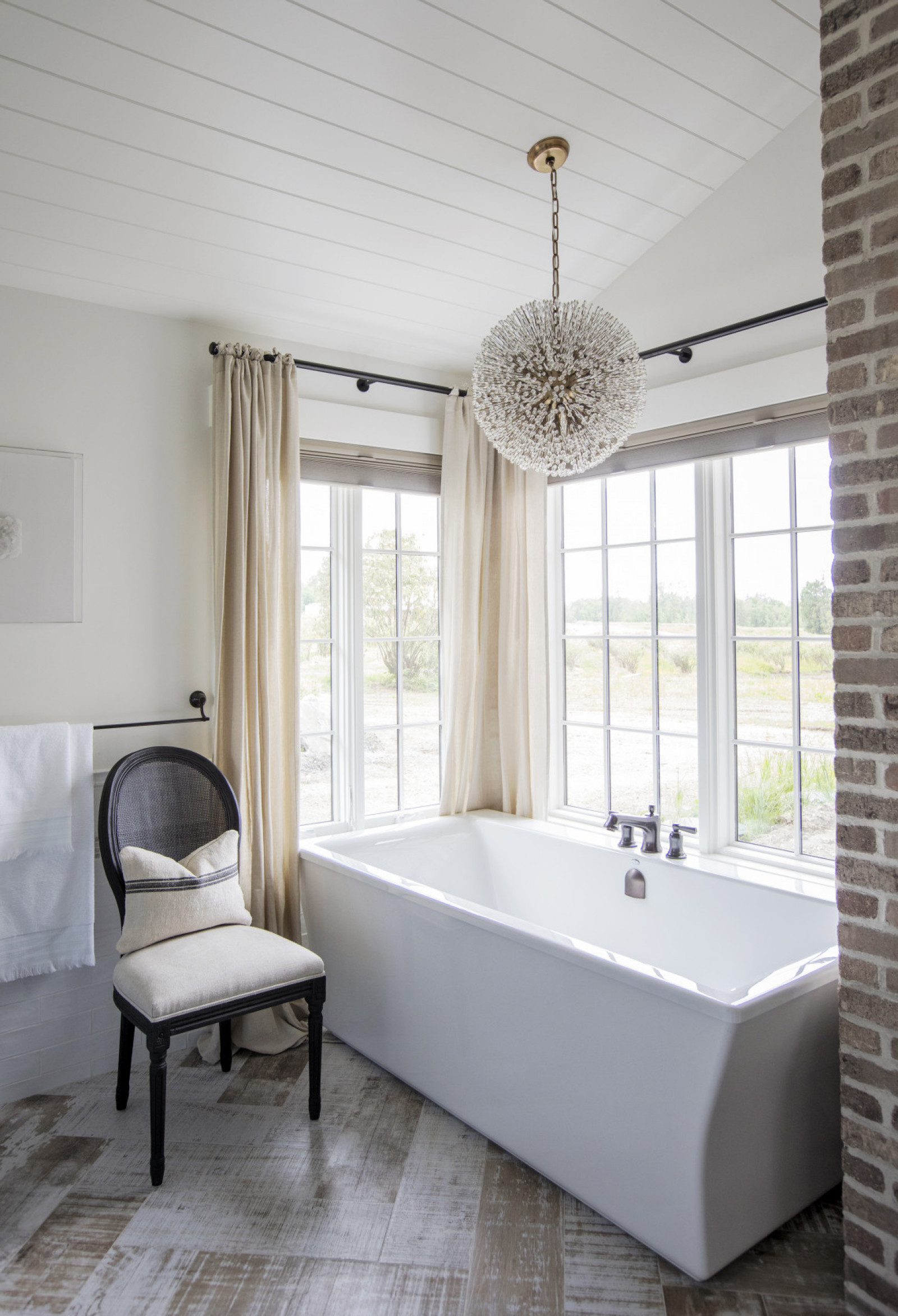 tub in front of window, upholstered chair with dark wood frame, whitewashed chevron floor, orb chandelier