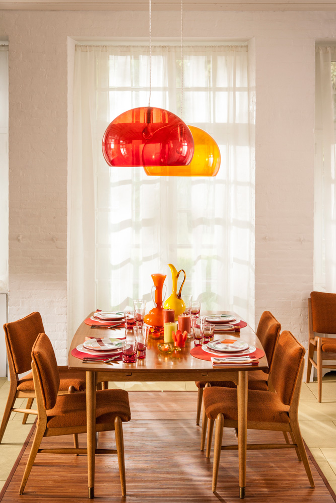 A dining room with a wooden dining set including chairs upholstered in orange textile. Red and orange lighting hangs overhead while red and orange dinnerware is arranged on the table.