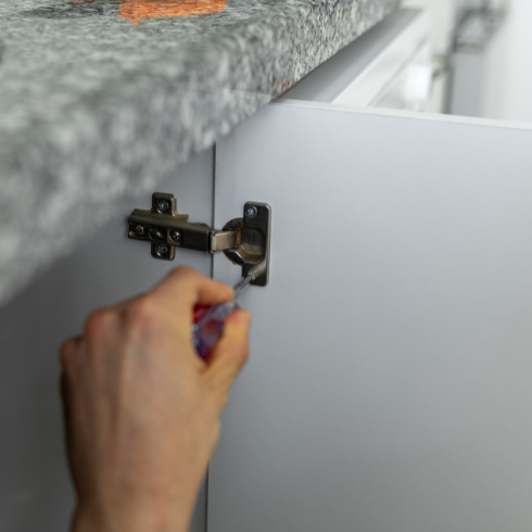 A closeup of hands fixing a metal hinge on the inside of a white cabinet.