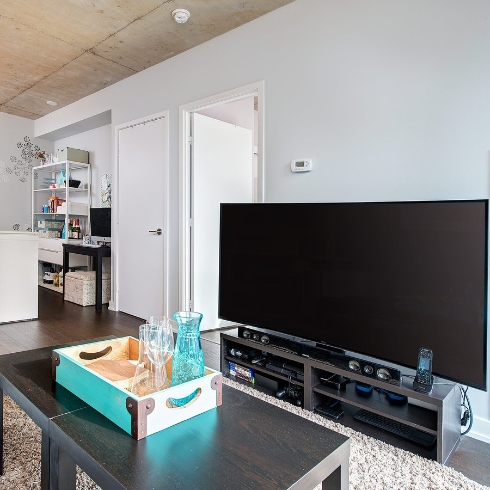 A condo living room with large flatscreen TV, a dark brown wooden coffee table and the kitchen in the background