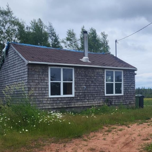 A small bungalow with brown shingle siding and roof, and two large windows