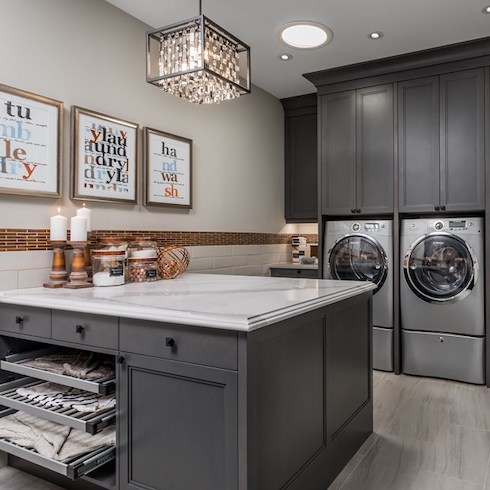 Spacious and fully functional laundry room with grey cabinets, a centre island with built-in drying racks, a crystal chandelier and large grey tile floors as part of a home reno trends story for Home Network