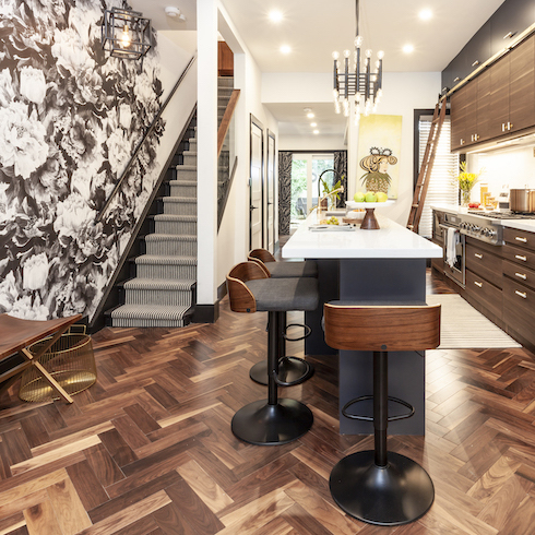 An opulent kitchen renovation by Save My Reno featuring a glass bannister and striped runner, a bold black-and-white floral wallpaper over the entire entry, dark herringbone parkay flooring, wood veneer bar stools and dark cabinets wall as part of a home reno trends story for Home Network