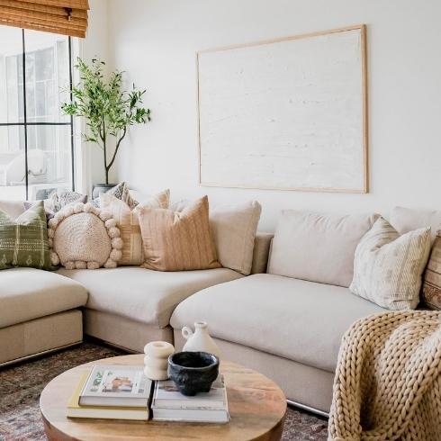 Large taupe corner sectional couch with piles of organic looking pillows in a white boho chic living room with large windows and a chunky knitted blanket as part of a home reno trends story for Home Network