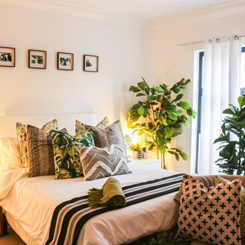 A bed with blankets and pillows in an assortment of patterns and colours with a large plant beside it