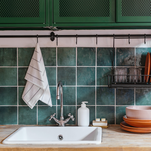 Interior design in kitchen with water faucet, white sink, black metal drying rack with ceramic plates and glass, wooden countertop and green tile on wall