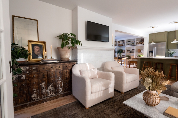 A living room wall with an apothecary cabinet, fireplace, TV and two large white armchairs