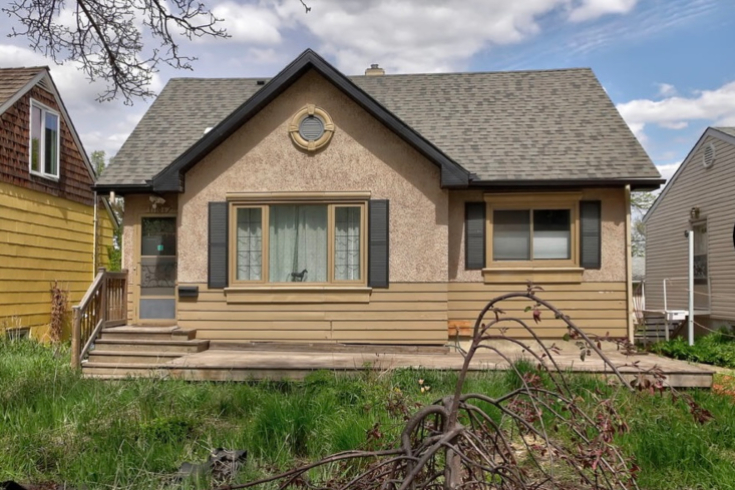 Exterior of a small home with painted stucco, dark grey shutters on two windows, and a main entrance to the side.