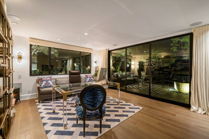 A study with one glass wall, another wall with large windows, a clear desk, blue chair and neutral sofa