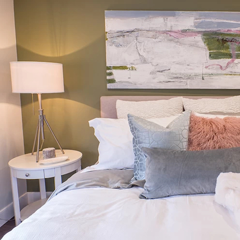 Beautiful bedroom with a sage green feature wall and large piece of art, a pink bed with white and grey bedding and a small round bedside table with a white lamp as featured on Home Network.