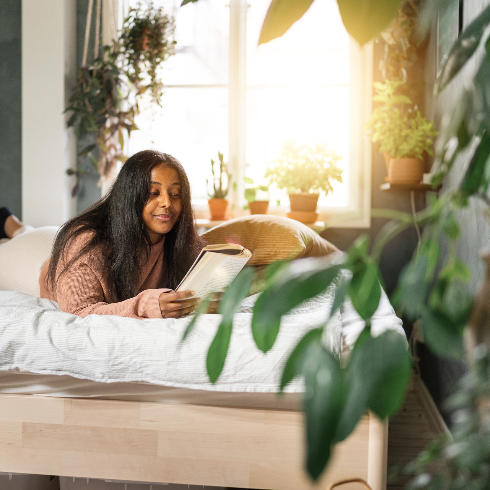 Woman is reading a paperback book at home. She is lying in her bed, surrounded by potted plants