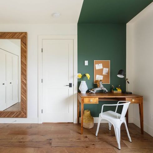 Home décor renovation designed by David Strongman of an office space with wide plank wood floors, a large wall mirror, white door, wooden desk, white desk chair, and a corkboard on a wall painted in emerald green as featured on Home Network