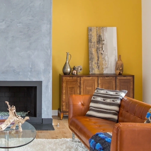 Chic living room with an orange leather couch, wooden credenza topped with vases and an abstract painting, grey fireplace, glass coffee table and walls painted in Dunn-Edwards Paints Honey Glow (DE5354)