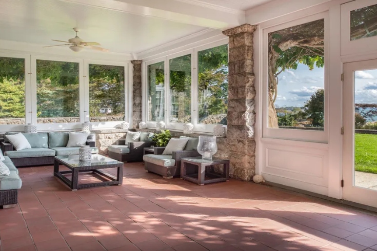 An enclosed porch area with stone walls, terra cotta tiled floor, large windows and a ceiling fan.