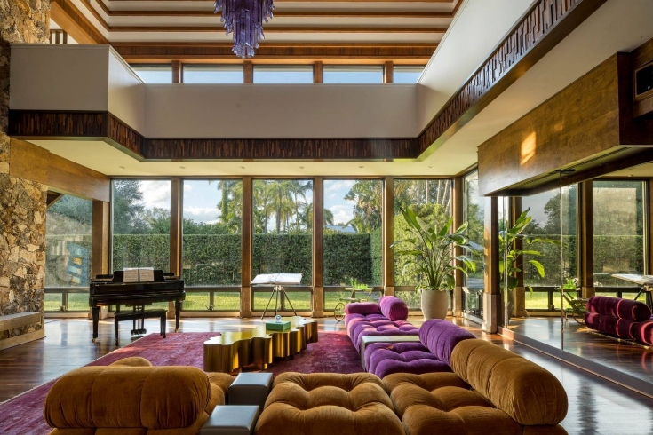 A living room with colourful sofas and chairs, floor-to-ceiling windows and a piano