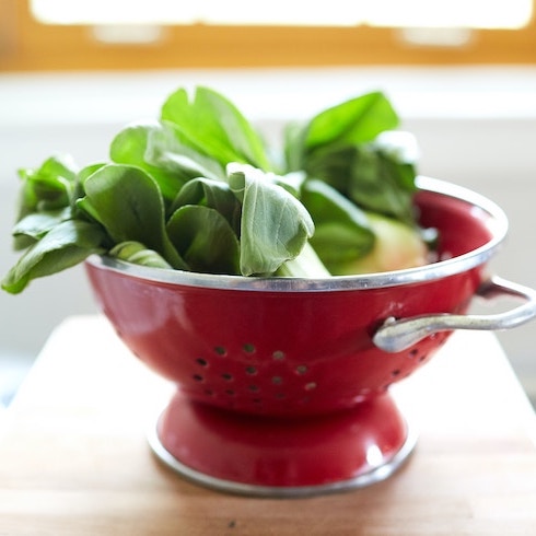 Green bok choy in a red strainer on top of a wood board