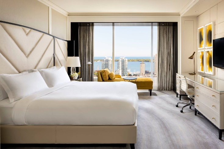 A hotel room with platform-style bed, grey carpeting, large window and framed sunflower art.