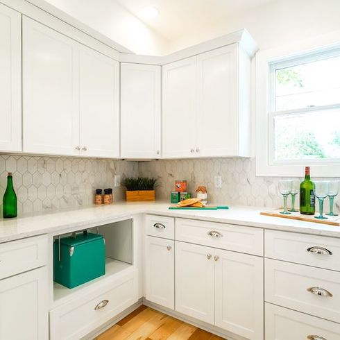 Roomy, pristine prep kitchen renovation from Masters of Flip stars Dave and Kortney Wilson for Home Network featuring white floor to ceiling cabinets, a perfectly placed window, and a white marble topped counter with green bottles, a cutting board, tea tins, wine glasses, spices and a potted plant