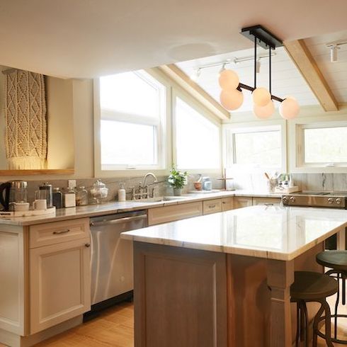 A bright white kitchen featuring new, energy-efficient windows, a multi globe pendant light over the kitchen island, white shaker style cabinets, white shiplat and exposed beam ceiling, and white marble countertops designed by Scott McGillivray for Home Network’s Scott’s Vacation House Rules kitchen