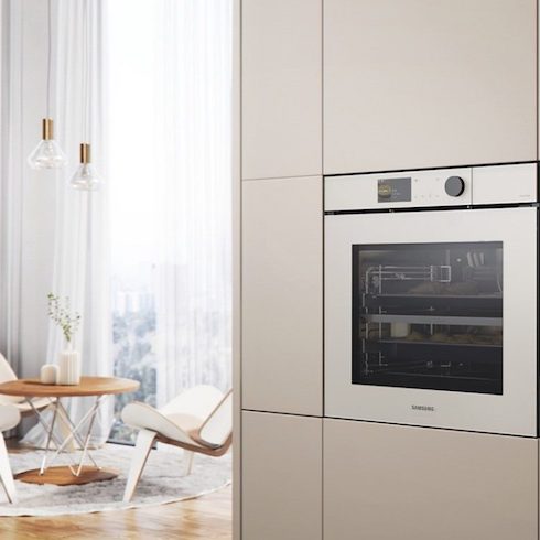 Samsung’s new AI oven, which uses AI tech to monitor the cooking process, is installed in a white panelled wall beside a chic sitting area with white bentwood armchairs, a white round rug, a round coffee table with two white vases on top, long gauzy drapes and two glass and good pendant lamps