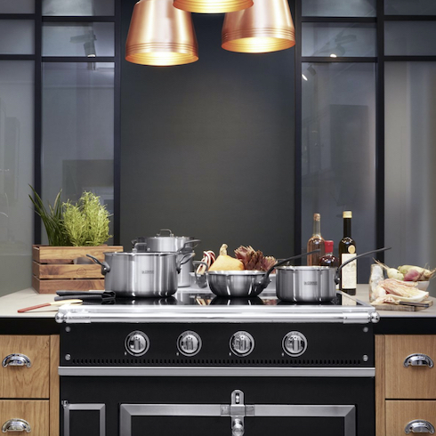 A black La Cornue Chateau induction cooktop is laden with magnetisable pots and surrounded by food, herbs and cooking utensils in a dark kitchen with wood faced cabinets and three large copper pendant lamps overhead