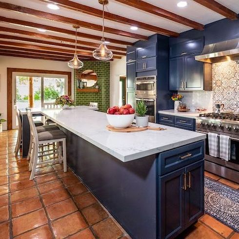 Beautiful kitchen in a Las Vegas rancher renovated by Jonathan and Drew Scott on Home Network’s Canada’s Property Brothers: Forever Home featuring a long T-shaped blue kitchen island, white quartz countertops, blue cabinets, exposed wood beams, large terracotta floor tiles, two glass pendant lamps, and a green brick fireplace in the corner