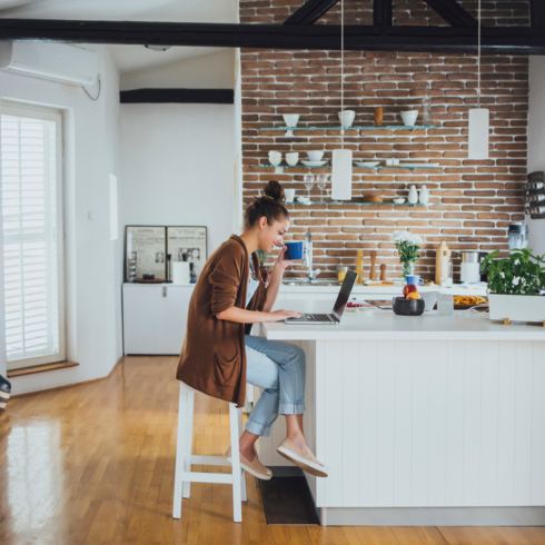 a young white millennial woman sitting at her laptop in a kitchen with a brick accent wall