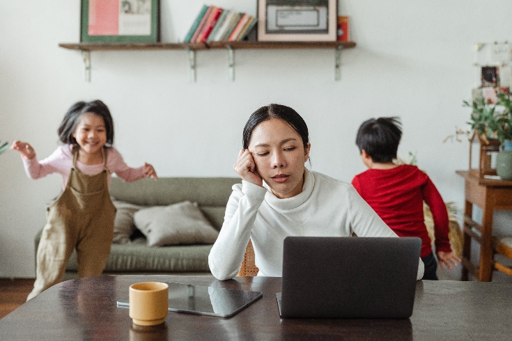 A stressed mother in front of a computer while her two kids run around in the background