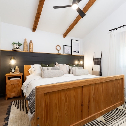 White and black bedroom with a black panelled headboard wall feature with two black wall sconces and an art shelf, a black and white rug, a blanket ladder, a wooden bedside table, a black ceiling fan and two exposed wooden ceiling beams