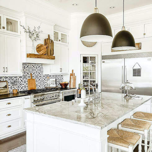 Two Maddox Black Dome Pendant lights designed by Blake Tovin of Tovin Design for Crate & Barrel featured in Home Network’s kitchen lights gallery in a modern farmhouse kitchen with a large centre island with marble countertop, three wicker stools, white cabinets, mosaic tile backsplash with a wood shelf above the stainless steel stove